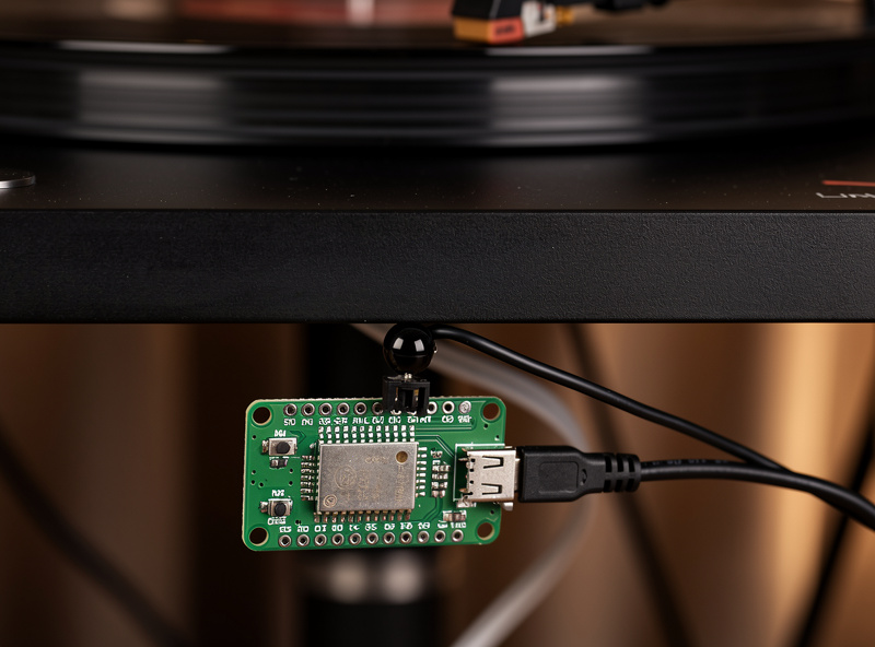 Close-up of a small ESP32-based sensor module mounted discreetly under a turntable plinth with a wire running to a smart plug, soft low light hi-fi rack scene