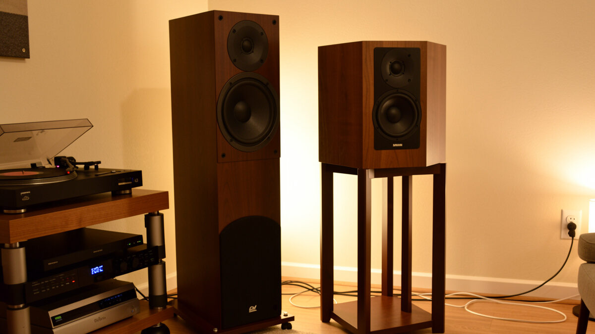 Floorstanding tower speaker beside a bookshelf speaker on a stand in a vinyl listening room