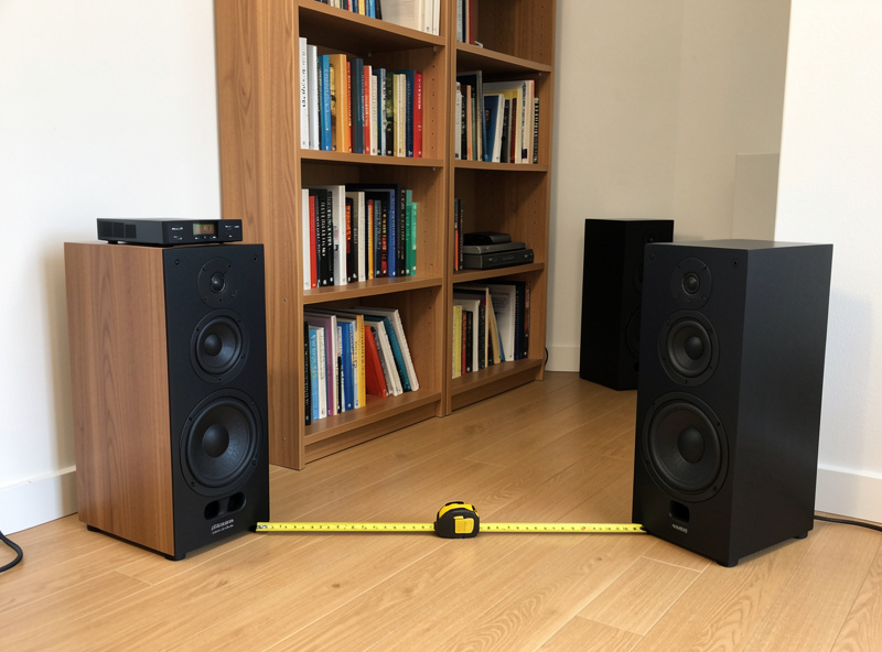 Bookshelf speakers pulled out from the front wall in a listening room with a measuring tape on the floor