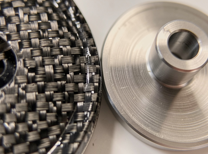 Macro close-up of the woven texture of a carbon fiber headshell next to a machined aluminum one