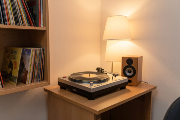 A compact turntable on a small desk in a cozy dorm room with records on a shelf and a bookshelf speaker
